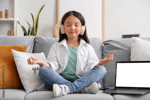 Little Asian girl in headphones with laptop meditating on sofa at home