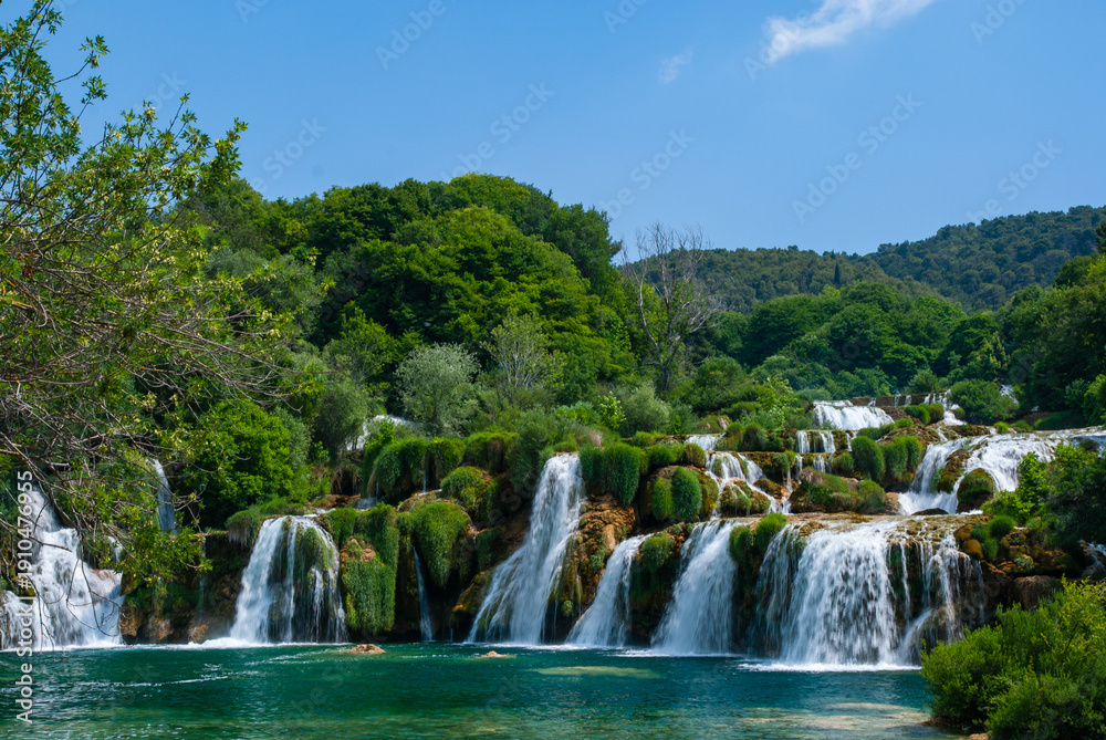 Naklejka premium Stunning series of cascading waterfalls surrounded by lush greenery, with clear blue water flowing over moss-covered rocks under bright sky. Krka National Park is of Croatia's natural treasures.