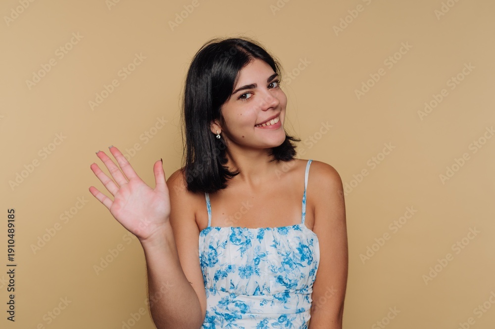 Obraz premium Smiling young woman waves cheerfully against a warm, neutral background during a sunny afternoon
