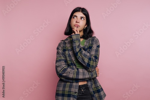 Thoughtful young woman in a plaid shirt poses against a soft pink backdrop, pondering life’s questions with a curious expression