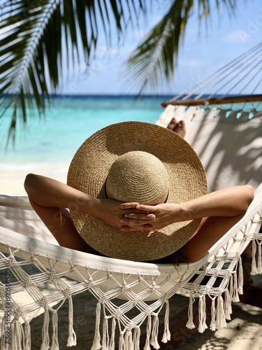 A woman is laying on a hammock on a beach, wearing a straw hat