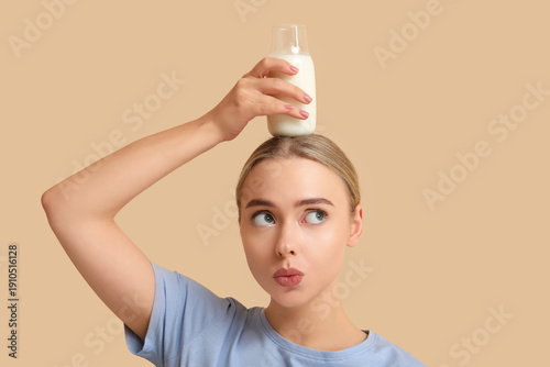 Pretty young woman with glass bottle of milk on her head against beige backgr...
