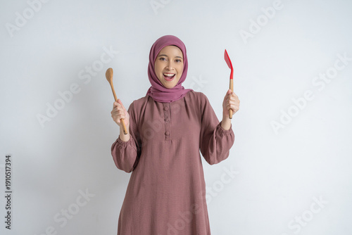 Tablou pe pânză A smiling woman in a pink hijab and modest dress stands against a light background, holding a wooden spoon and a red spatula, conveying warm, home cooking and kitchen vibes