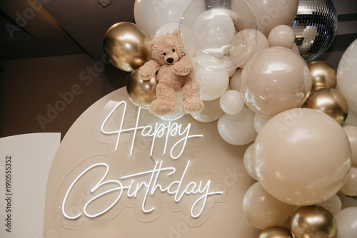 A photo backdrop for a birthday photo shoot decorated with balloons, neon lettering, and soft toys
