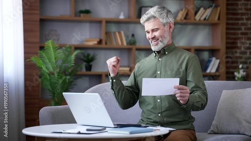 Happy man with gray hair sitting at home office holding document and looking at laptop with excitement. Male celebrating after receiving positive financial news, social benefits, or tax refund online
