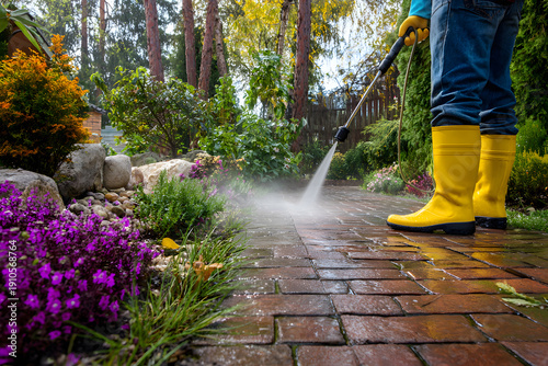 Person pressure washing brick pathway in garden with yellow boots