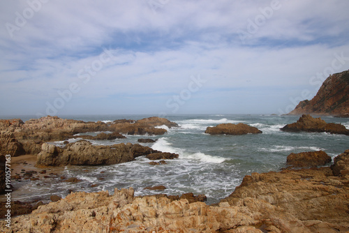 Rocky coastal bay with waves breaking between stone formations near Knysna, South Africa, photographed in daylight under a lightly clouded sky.