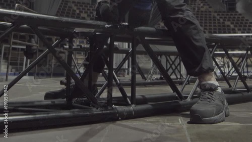Man adjusts base of long truss bar. Worker stands firmly on arena floor balancing lighting support frame before full rig assembly begins