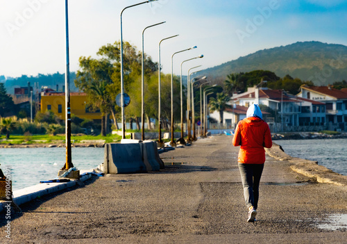 Fotografi Rear view of a person in a red jacket and hoodie walking along a coastal road in an Aegean town, peaceful morning stroll by the sea with mountains and street lamps