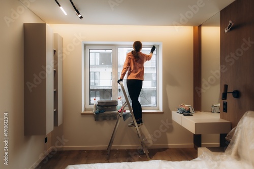 Woman cleaning window using ladder in modern apartment