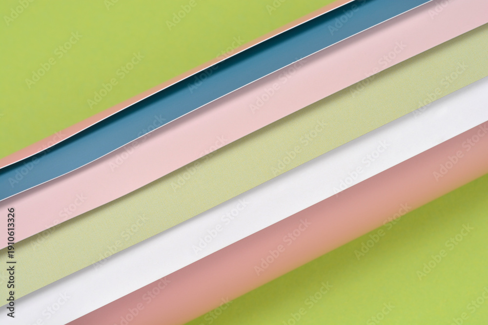 Fototapeta premium Top view of several overlapping layers of pastel colored plastic sheets in pink blue and white arranged diagonally over a solid green surface for creative background