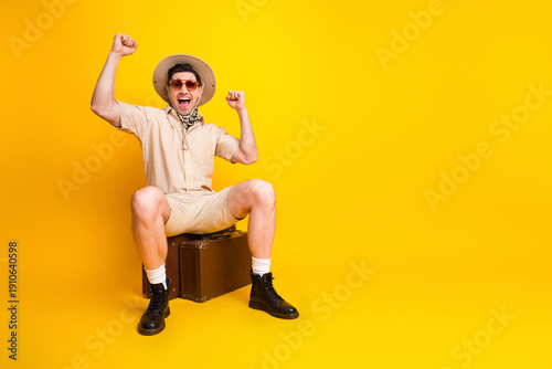Fotografija Cheerful adventurer in safari outfit sits on a suitcase against a bright yellow