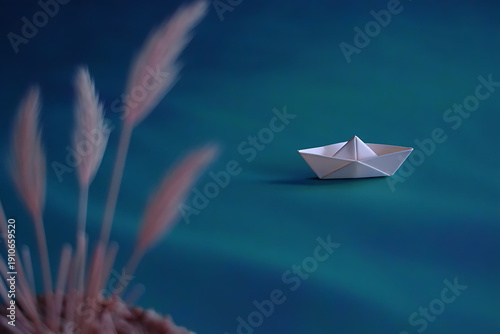 A small white paper boat floats on a calm blue surface next to blurred plants