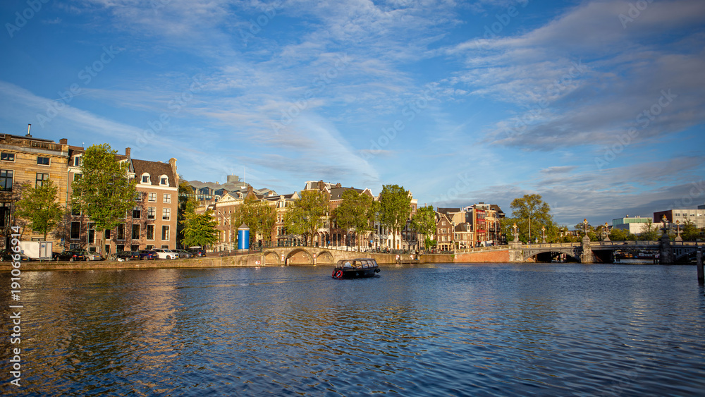 Obraz premium Wide Amsterdam canal with bridge and skyline. Panoramic Amsterdam canal scene with historic buildings, trees, small boat on the water, and a bridge beneath a bright blue sky.