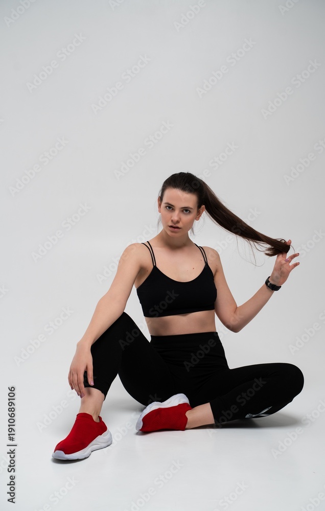 Fototapeta premium Young fitness woman in black sports bra and leggings sitting on a studio floor, pausing from workout with strong body language, focusing on wellness and healthy lifestyle