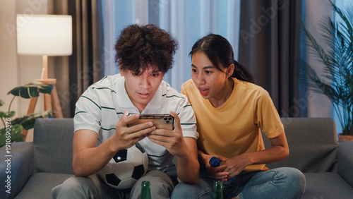 Asian new marriage couple sit on sofa excited cheering watching football sport game on mobile phone in living room at home. Young couple sport fans hold football watching online score on smartphone.