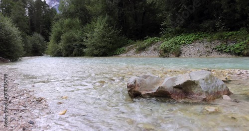 Alpine stream swirling around a large boulder in italian alps video, clear turquoise water runs over pebbles beside dense forest in soft overcast light