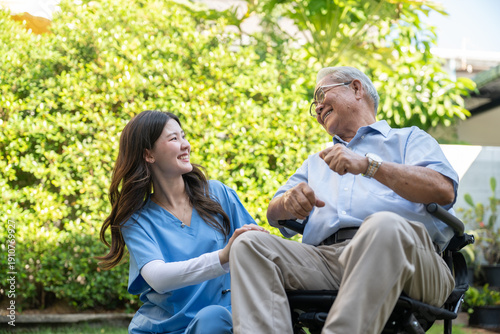 Smiling elderly patient sitting in wheelchair with professional nurse assistant taking care outdoors at nursing home facility.