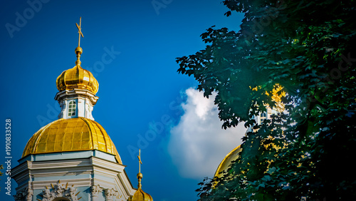Golden domes of an Orthodox church against a vibrant blue sky. Lush green trees frame the architecture. A beautiful religious landmark