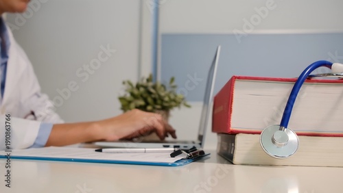 Medical professional typing on laptop at desk with medical books and stethoscope, working on healthcare research, telemedicine, or patient documentation