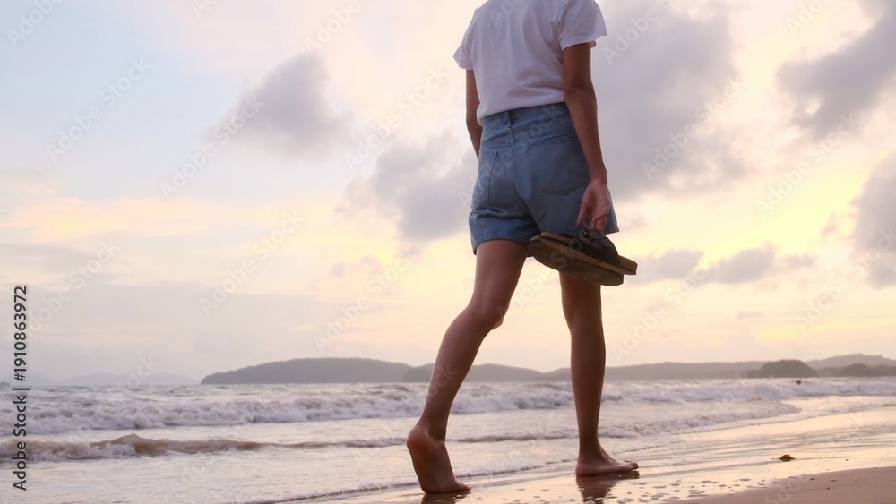 Fototapeta premium Young woman walking barefoot along a sandy beach shoreline at sunset, carrying sandals in hand, enjoying freedom and peace on a mindful solo ocean stroll