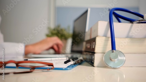 Schilderij op canvas Medical professional typing on laptop at desk with stethoscope, stack of textboo