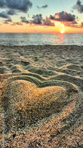 Heart shape drawn in sand during sunset at the beach by the ocean with waves and clouds in the background