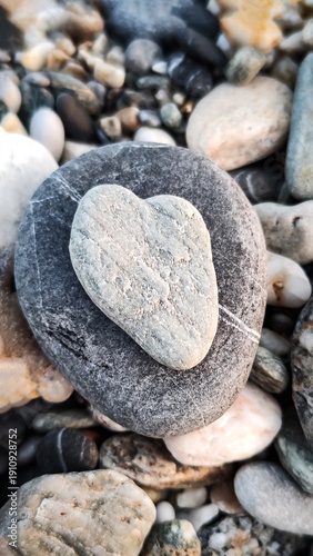 Heart-shaped stone sits on a larger rock among other smooth stones by the shore during daylight along a beach setting