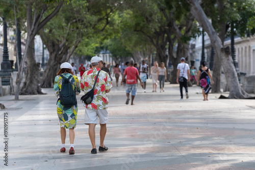 Older  couple of tourists in bright clothes and hats walking on Prado boulevard in the city center of Havana, Cuba. Selective focus, blurred background. Vacation, travel, tourism concept. 
