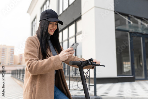 Фотография oman in a beige coat on a electric scooter uses a smartphone against the background of office buildings