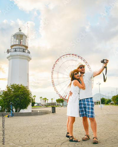 Young couple in love smiling and taking selfie at Batumi Boulevard with iconic lighthouse and Ferris wheel during golden hour. Happy couple Holiday taking photo with popular landmarks