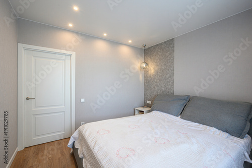 A minimalist bedroom with a double bed, grey pillows, white quilt, light grey walls, recessed ceiling lights, a pendant lamp, white door, and light wood flooring