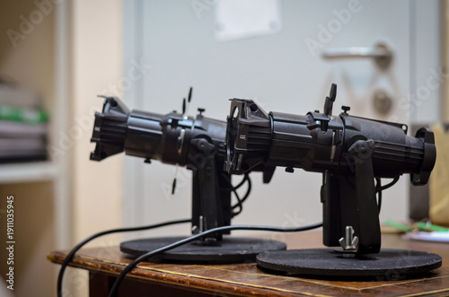 Pair of professional stage spotlights on stands with lenses and adjustment controls, positioned indoors on a table with shallow depth of field.