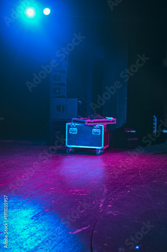 Moody stage scene with neon blue and magenta lighting illuminating audio equipment case and speakers, creating a dramatic concert or club atmosphere.