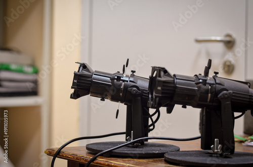 Pair of professional stage spotlights on stands with lenses and adjustment controls, positioned indoors on a table with shallow depth of field.