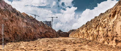 Workers are preparing the construction site by digging deep trenches for a new building as cranes loom over the area under a cloudy sky
