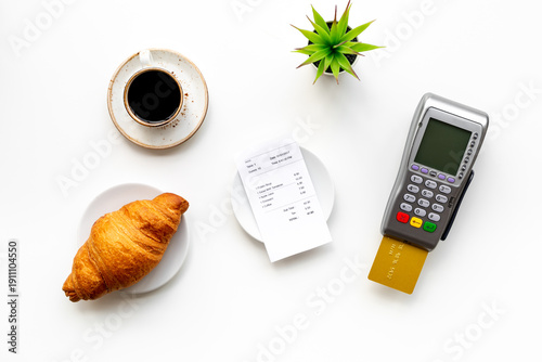 Paying the bill by pos terminal in cafe or restaurant. Cup of black coffee and croissant, payment wireless machine and bill on white background, top view