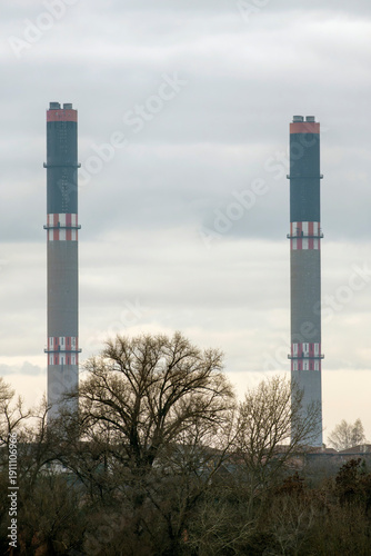 Tall chimneys stand against a cloudy sky with bare trees in the foreground near an industrial area at dusk