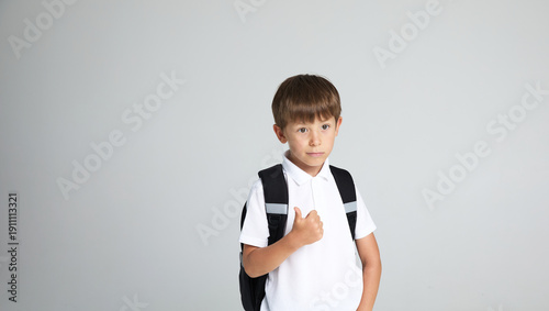 Horizontal view of a amusing little boy with a backpack on his back, holding hand up, isolated on gray background. Copy space.