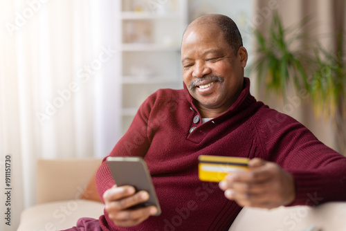 Middle aged black man using smartphone and holding credit card for online shopping at home, secure payment and digital banking concept in cozy living room environment