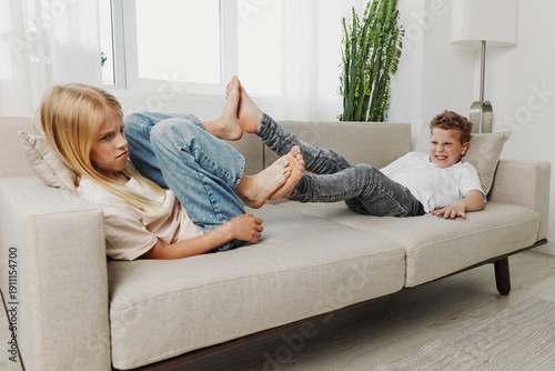 Fotografie Two children lying on a sofa, playfully interacting with their feet, showing sibling rivalry and playful mood in a bright living room
