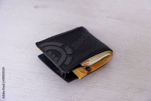 A black leather wallet is closed and visibly bulging with the large number of 50-euro notes inside. Although not fully visible, some orange edges of the notes protrude slightly, 