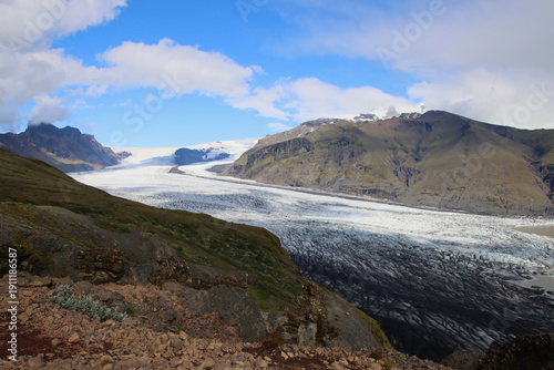 Skaftafellsjokull glacier tongue flowing from the Vatnajokull ice cap in Skaftafell, Southeast Iceland, surrounded by volcanic mountains under partly cloudy sky.