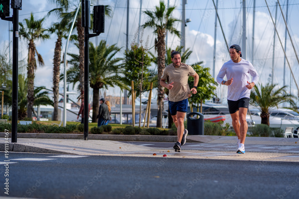 Obraz premium Two men jogging together along a sunny marina promenade with palm trees and boats, enjoying an active urban lifestyle focused on fitness, friendship and outdoor wellbeing
