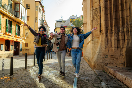 Fotografia Three happy young adult friends holding hands and running with arms open, enjoyi