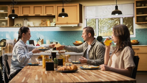 Obraz na plátne Parents and kids sitting together at breakfast table being served by the mother, serving scrambled eggs with bacon and goods