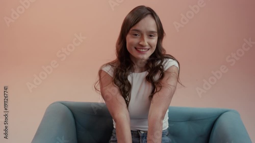 Medium shot shows a young woman with brown hair sitting in a turquoise armchair. She is wearing a white top and touching her face with both hands against a uniform peach background. 4K.