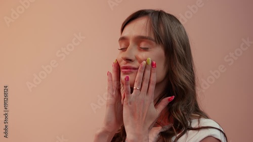 Close-up shot of a young woman with brown hair and multi-colored nails. She touches her face with her eyes closed, against a peach, uniform studio background. 4K.