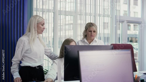 Medium shot of two Caucasian female employees supervising a female colleague working on a computer. The women in the back are wearing white shirts, against a bright window backdrop. 4K.