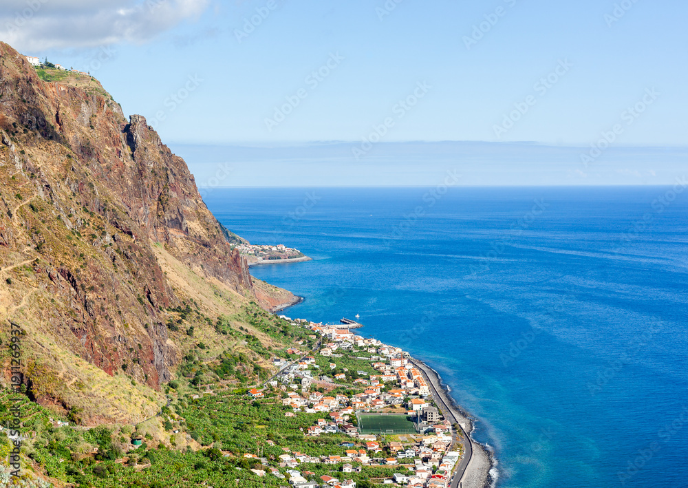 Fototapeta premium Madeira cliffside village. Steep rock face towers above terraced homes and green plots. Bright blue ocean fills the right side of the frame. A curving shore road runs along the coast.